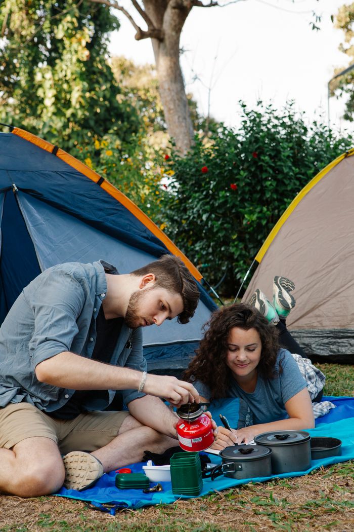Concentrated young male hiker in casual clothes switching on portable gas stove while sitting near tent with girlfriend during camping in green woods